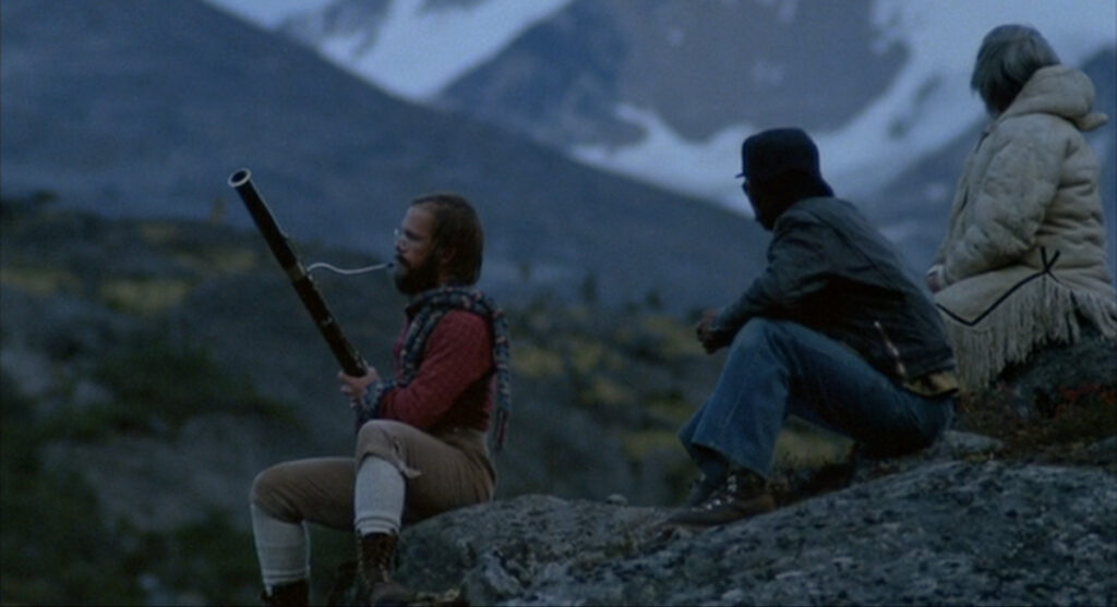 Tyler sits on a rock and plays bassoon while Mike and Ootek look on